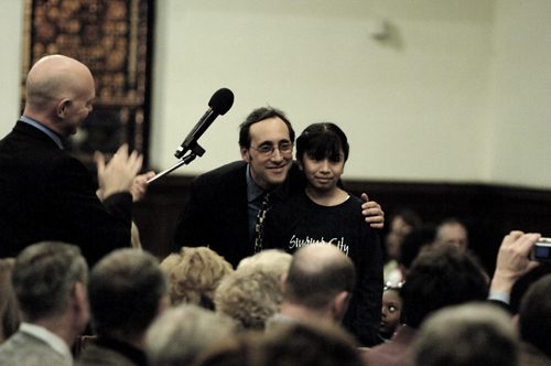 Andrew Bleckner on stage with a performer and a speaker at a microphone, in front of an audience